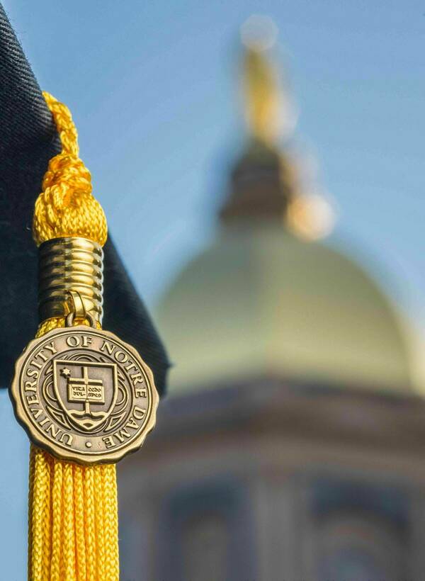 Graduation cap with gold tassel and Notre Dame emblem with a blurred golden dome in the background.