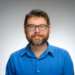 Headshot of a man with short brown hair, glasses, and a beard. He wears a bright blue, button-down collared shirt. He has a slight smile.