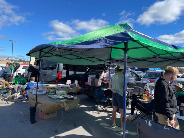 Under a green and blue canopy tent, Notre Dame fans tailgate. People in green and blue apparel gather around a table with food containers, next to parked vehicles on a sunny day. The Joyce Center sign is visible in the background.