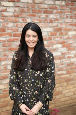 Smiling woman with long dark hair wears a black floral dress, hands clasped, against a brick wall.